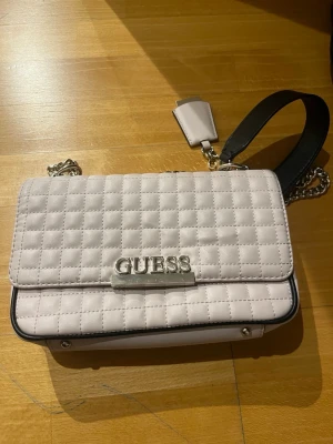 Ljusrosa quiltad axelväska från Guess - Snygg ljusrosa axelväska från Guess med quiltat mönster och silverfärgad logotyp framtill. Väskan har kedjerem med svart axelparti och detaljer i silver. Perfekt för att lyfta din outfit med en trendig touch. Äkta