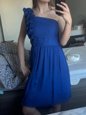 Blå one shoulder klänning med volang - Snygg blå klänning med one shoulder-design och volangdetaljer längs axelbandet. Storlek 2 vilket motsvarar M men som referens brukar jag ha xs/s