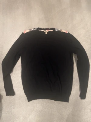 Svart stickad tröja från Burberry - Svart stickad tröja från Burberry med klassiskt rutmönster i beige, rött och grått på axlarna. Tröjan har rund halsringning och långa ärmar. Perfekt för dig som gillar stilrena plagg med en twist. Där det står storlek är L fast sitter som S samt den är lite sönder 