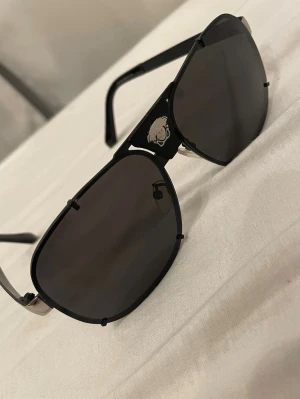 Svarta pilot-solglasögon - Snygga svarta solglasögon i klassisk pilotmodell med mörka linser. Bågarna är i metall med silverfärgade detaljer och sidorna samt Medusa-emblem framtill. Perfekt accessoar för en trendig look.