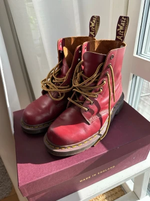 Vinröda Dr. Martens boots i läder - Säljer ett par vinröda Dr. Martens boots i äkta läder med klassiska gula sömmar och grov sula. Skorna har högt skaft, bruna och gula snören samt tydliga Dr. Martens-taggar baktill. Tillverkade i England och perfekta för en edgy stil. Model: vintage, stl: 39. knappt använda, ca en månad pga. Skada/olycka.