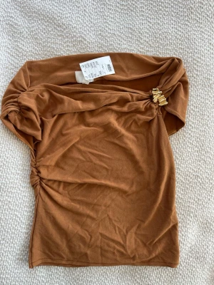 Brun offshoulder topp med gulddetalj - Snygg brun offshoulder topp med guldfärgade detaljer vid axeln. Nyskick med lappen kvar 