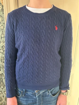 Mörkblå kabelstickad tröja Polo Ralph Lauren - Kabelstickad mörkblå tröja från Polo Ralph Lauren med rund halsringning och röd broderad logga på bröstet. Tröjan har långa ärmar och ribbade muddar vid ärmslut och nederkant. Perfekt till jeans för en klassisk look.