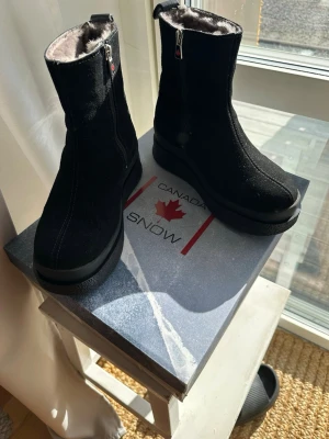 Svarta vinterstövlar från Canada Snow - Säljer ett par svarta vinterstövlar från Canada Snow med dragkedja på sidan och mjukt foder inuti. Ovandelen är i mocka och baksidan har en snygg detalj i lack. Perfekta för kalla dagar och riktigt sköna att ha på sig. Använda två veckor. 