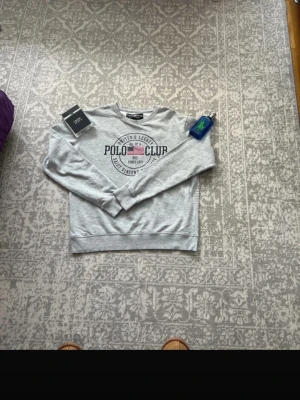 Grå sweatshirt från Polo Club - Snygg grå sweatshirt från Polo Club med tryck framtill och rund hals. Tröjan har långa ärmar och klassisk passform, perfekt för en avslappnad stil. Mjukt material som känns skönt mot huden.