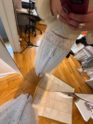 Vit kjol med småmönster - Vit kjol med lös och vid passform, dekorerade med små rosa och grå prickar. Hög smockad midja för extra komfort och ett mjukt, tunt material som känns skönt mot huden. Perfekt för en sommardag.