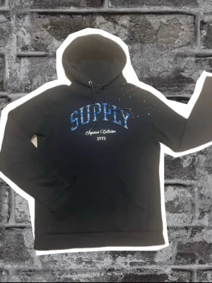 Svart hoodie Supply & Demand XS - Svart hoodie från Supply & Demand med blått SUPPLY-tryck och vita detaljer på bröstet. Klassisk känguruficka och dragsko i huvan. Materialet är mjukt och bekvämt, perfekt för en avslappnad streetstyle-look.