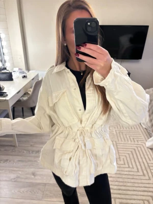 Vårjacka - Säljer en snygg vit vårjacka med stora bröstfickor och dragsko i midjan. Jackan har klassisk skjortkrage, knäppning framtill och är tillverkad i ett lätt tyg som ger en avslappnad look. Perfekt att slänga över en t-shirt för en cool stil.