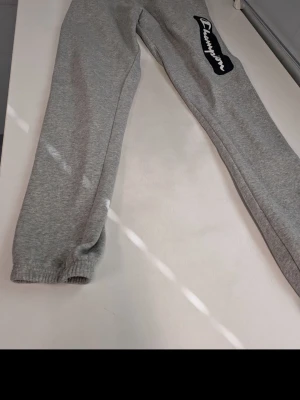 Gråa mjukisbyxor från Champion - Gråa mjukisbyxor från Champion med stort logotryck längs ena benet. Byxorna har resår i bensluten och är tillverkade i mjukt material, perfekt för chill dagar. Passar dig som gillar sportig och avslappnad stil.