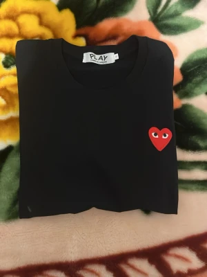 Svart Comme des Garçons Play t-shirt - Svart t-shirt från Comme des Garçons Play med klassisk rund hals och korta ärmar. På bröstet sitter det ikoniska röda hjärtat med ögon. Perfekt för dig som gillar stilrena plagg med en twist.