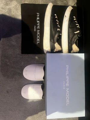 Philippe Model skor - Svarta Philippe Model skor i läder med vita detaljer och vita snören. Klassisk låg modell med vit sula och diskret logga på sidan. På bilderna ser du äkthetsbevis på originalkartong och skopåsar. Perfekta för dig som gillar stilrena och tidlösa sneakers.