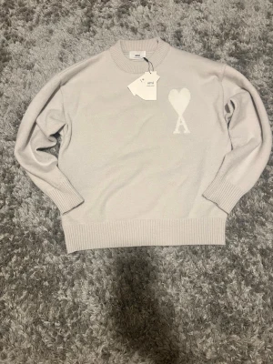  Ami Paris sweater  - Snygg ljusgrått stickad tröja från Ami med rund halsringning och ribbade muddar. På bröstet finns det ikoniska Ami-hjärtat med bokstaven A i vitt. Perfekt för en stilren och trendig look.