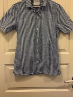 Randig blå skjorta från Primark - Snygg kortärmad skjorta från Primark i blått och vitt med smala ränder. Skjortan har klassisk krage, knäppning framtill och är tillverkad i ett lätt linnematerial som passar perfekt till varma dagar. Storlek XS, men passar S
