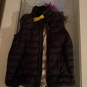 Svart dunväst från Parajumpers - Säljer en svart quiltad dunväst från Parajumpers med gul detalj vid kragen och dragkedja framtill. Västen har två fickor med dragkedja och en hög krage. Insidan är ljusbeige och västen har klassisk Parajumpers-logga på ryggen. Har ingen dragsedja