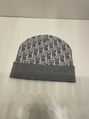 Grå Dior mössa med logotypmönster - Snygg grå mössa från Dior med ikoniskt vitt logotypmönster över hela ytan. Mössan har en vikt kant och är tillverkad i mjukt stickat material som ger en clean och trendig look. Perfekt accessoar för dig som vill ha något exklusivt och stilrent.