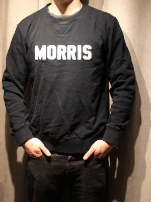 Svart sweatshirt från Morris - Cool svart sweatshirt från Morris med vit broderad logga på bröstet. Tröjan har rund halsringning, ribbade muddar och avslappnad passform. Perfekt för en avslappnad och stilren look. Modellen är 180cm