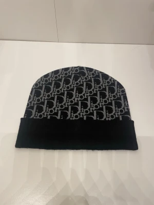 Svart och grå Dior mössa - Snygg svart mössa från Dior med grått logomönster över hela ytan. Klassisk form med uppvikt kant och stickad känsla. Perfekt accessoar för att lyfta din streetwear-look.