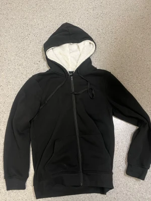 Svart hoodie med dragkedja och foder - Mysig svart hoodie med dragkedja och vit fluffig fleece på insidan av huvan. Klassisk modell med fickor framtill och justerbar dragsko i huvan. Perfekt för chill dagar eller när du vill ha något bekvämt och stilrent.