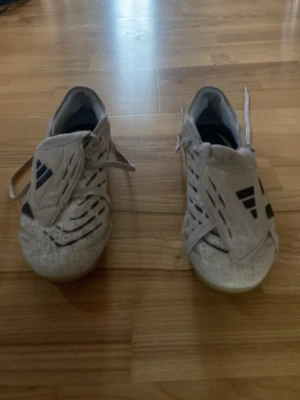 Vita Adidas fotbollsskor med svarta detaljer  - Säljer ett par vita fotbollsskor från Adidas med svarta detaljer och snörning. Skorna har en modern design med mönstrad ovandel och klassisk Adidas-logga på tungan. Perfekta för dig som vill sticka ut på planen.