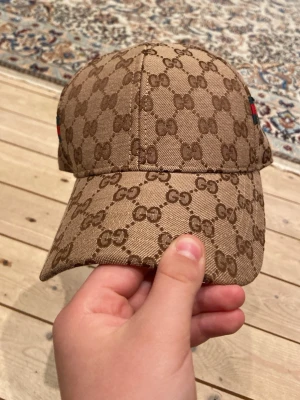 Beige keps med GG-mönster - Säljer en beige keps från  med det klassiska GG-monogrammet över hela kepsen. På sidan finns en ikonisk grön och röd rand. Kepsen har böjd skärm och justerbar rem bak. Materialet är canvas med exklusiv känsla.