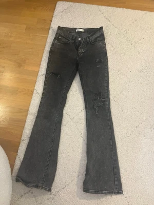 Gråa bootcut jeans med slitningar - Säljer ett par gråa bootcut jeans med coola slitningar på benen. Jeansen har klassisk femficksdesign och är tillverkade i denim. Passformen är utsvängd nedtill och de har en snygg vintage-look.