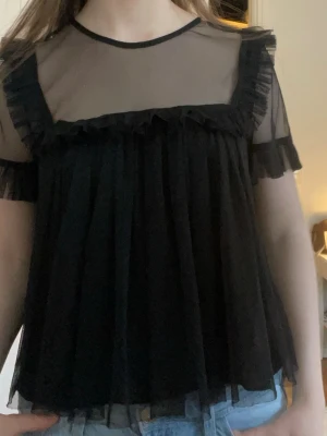 Svart transparent volangblus - Säljer en svart blus med transparent upptill och volangdetaljer längs bröstet och ärmsluten. Blusen har en lös passform och korta ärmar! 
