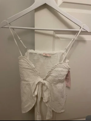 Vit linnetopp från Nelly med knyt - Supersöt vit linnetopp från Nelly med smock över bysten, tunna justerbara axelband och knyt framtill. Toppen är ärmlös och har en luftig känsla, perfekt för varma dagar. Materialet är linneblandning som ger en fräsch och somrig vibe.