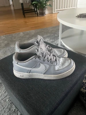 Nike Air Force 1 grå sneakers - Säljer ett par klassiska Nike Air Force 1 sneakers i ljusgrått läder med vit sula och perforerad tå. Skorna har den ikoniska swoosh-loggan på sidan och AIR-märkning på sulan. Perfekt för dig som gillar streetstyle och vill ha ett tidlöst sneaker-utseende.