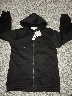 Cdg play hoodie  - Svart hoodie med dragkedja från Comme des Garçons Play. Framsidan har en liten hjärtlogga med ögon på bröstet och baksidan har en stor version av samma logga. Mjuk bomullskänsla och två fickor framtill. Perfekt för en stilren och cool look.