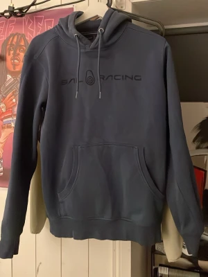 Blå Sail Racing hoodie med huva - Säljer en blå hoodie från Sail Racing med svart logga på bröstet. Tröjan har en stor ficka framtill, dragsko i huvan och ribbade muddar. Perfekt för dig som gillar sportig och avslappnad stil. Materialet känns mjukt och bekvämt.