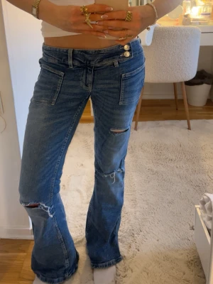 Blå slitna bootcut jeans med låg midja - Säljer ett par blå bootcut jeans med låg midja och slitningar på benen. Jeansen har dubbla knappar i linningen, stora framfickor och en avslappnad passform. Säljer då dem blivit för stora💖