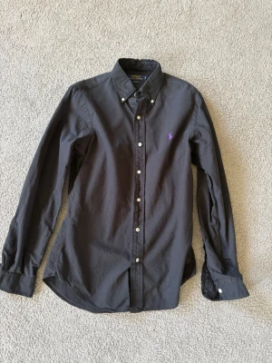 Mörkgrå skjorta från Polo Ralph Lauren - Klassisk mörkgrå skjorta från Polo Ralph Lauren med lila broderad logga på bröstet. Skjortan har lång ärm, knappar framtill och button-down krage. Tillverkad i mjuk bomull för en bekväm känsla och snygg passform.