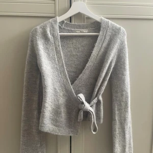 Stickad kofta gina - Säljer en ljusgrå omlottkofta i stickad kvalitet. Koftan har långa ärmar, knytband i sidan och ribbade muddar. Den är knappt använd och helt slutsåld på Gina Tricots hemsida💕