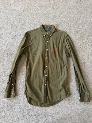 Olivgrön skjorta från Ralph Lauren - Säljer en olivgrön skjorta från Ralph Lauren med klassisk krage och långa ärmar. Skjortan har knappar framtill och en röd broderad logga på bröstet. Tillverkad i mjuk bomull som känns skön mot huden. Perfekt för dig som gillar stilrena plagg med ikonisk detalj.