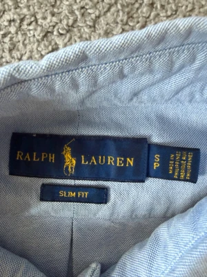 Ljusblå skjorta från Ralph Lauren - Klassisk ljusblå skjorta från Ralph Lauren i slim fit-modell. Skjortan har knappar framtill, button-down krage och den ikoniska broderade loggan på bröstet. Perfekt för en stilren och tidlös look.