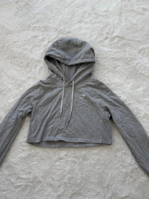 Grå cropped hoodie från Gymshark - Grå cropped hoodie från Gymshark med huva och dragsnören. I storlek S. Materialet är mjukt och stretchigt bomullsblandat tyg.