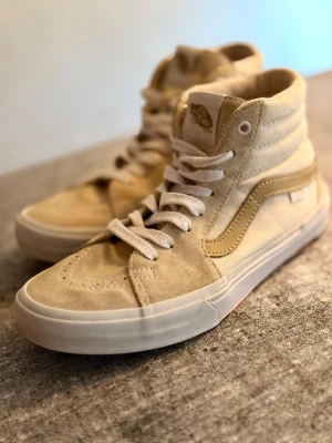 Beige Vans Sk8-Hi sneakers - Snygga beige Vans Sk8-Hi sneakers med klassisk vit sula och vit sidestripe. Skorna har högt skaft, vita skosnören och detaljer i mocka och canvas. Unik lotusblomma-detalj på hälen och Vans Off The Wall-logga baktill. Perfekta för en stilren streetlook.
