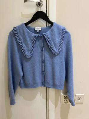 Ljusblå kofta med volangkrage I 💯 ull  - Supersöt ljusblå kofta från Others Stories med stora knappar framtill och en unik volangprydd krage. Koftan är kortare i modellen och har långa ärmar. Perfekt för dig som vill ha en gullig och trendig look.