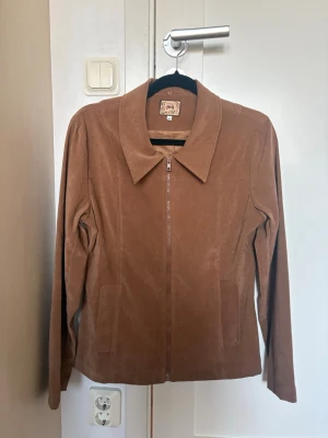 Brun jacka med utbytbar krage - Snygg brun jacka från Joy med rak passform och dragkedja framtill. Jackan kan bytas ut mot en mjuk, mörkbrun fuskpäls-krage för extra stil. Perfekt för dig som gillar att variera din look.