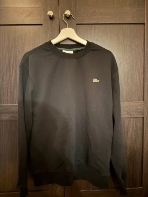 Svart sweatshirt från Lacoste - Klassisk svart sweatshirt från Lacoste med rund halsringning och den ikoniska gröna krokodilloggan broderad på bröstet. Tröjan har ribbade muddar vid ärmslut och nederkant.                                                                            Tröjan är helt ny. Råkade tvätta den i för hög värme och den blev lite för liten har använts 1 gång.