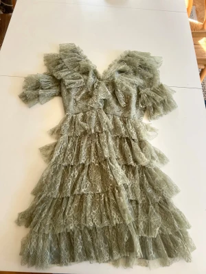 Grön volangklänning i spets - Supersöt kort grön klänning med flera lager volanger och spetsdetaljer. Klänningen har v-ringning, korta ärmar och är tillverkad i ett tunt, spetsigt material som ger en romantisk vibe. Perfekt för dig som gillar statement-plagg och vill sticka ut.