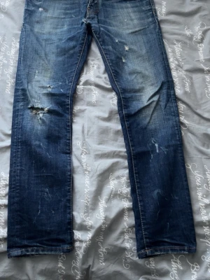 Dsq2 jeans herr - Säljer ett par blå jeans från dsquared2 med slitningar och hål för en riktigt cool och avslappnad look. Jeansen har raka ben, klassisk femficksmodell och knappgylf. Perfekta för dig som gillar en streetwear-stil.