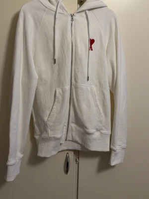 Vit hoodie med rött hjärta från Ami - Vit hoodie med dragkedja från Ami, prydd med det ikoniska röda hjärtat och bokstaven A broderat på bröstet. Hoodien har huva med dragsko, två fickor framtill och ribbade muddar. Tillverkad i mjuk bomull för en skön och avslappnad look. Den har storlek M men funkar oxå som L