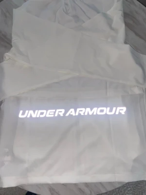 Vit Wind braker från Under Armour - Snygg vit hoodie från Under Armour med dragkedja och huva. Tillverkad i ett tekniskt material med UA Storm-teknologi som stöter bort vatten. Logga på bröstet och stor reflekterande text på ryggen. Perfekt för träning eller chill.