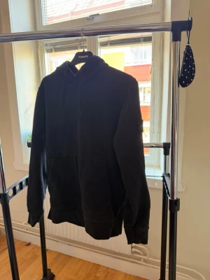 Svart hoodie från Stone Island - Svart hoodie från Stone Island med klassisk huva och känguruficka framtill. Ikonisk logotyp-patch på vänster ärm. Mjuk bomullskvalitet och avslappnad passform, perfekt för en streetwear-look.