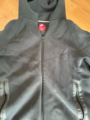 Svart Nike hoodie med dragkedja - Snygg svart hoodie från Nike med hel dragkedja framtill och två fickor med dragkedja. Klassisk Nike-logga på bröstet och huva för extra stilpoäng. Perfekt för en sportig och avslappnad look.