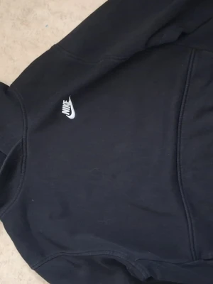 Svart Nike hoodie med logga - Svart hoodie från Nike med klassisk vit broderad logga på bröstet. Hoodien har huva och en stor magficka framtill. Tillverkad i mjukt bomullsmaterial och har ribbade muddar vid ärmar och nederkant. Perfekt för en avslappnad och sportig stil.