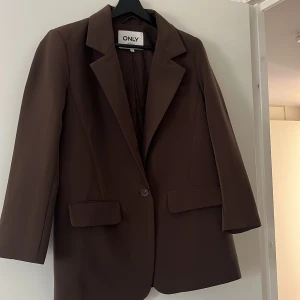 Brun kavaj från ONLY - Stilren brun kavaj från ONLY med klassisk krage och en knapp framtill. Kavajen har två fickor med lock och raka ärmar. Perfekt för en trendig och avslappnad look.