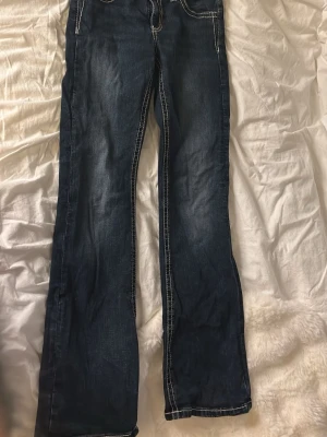 Mörkblå bootcut jeans med brodyr - Säljer ett par mörkblå bootcut jeans med kontrastsömmar och snygg broderad design på bakfickorna. Jeansen har låg midja och klassisk femficksmodell. Perfekta för dig som gillar en avslappnad men ändå trendig stil.