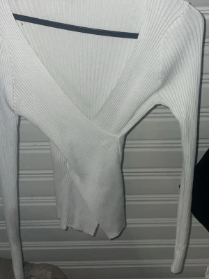 Vit ribbad omlottopp - Snygg vit omlottopp med ribbad struktur och djup v-ringning. Långärmad modell som sitter tight och framhäver figuren. Perfekt att styla med jeans eller kjol för en trendig look.